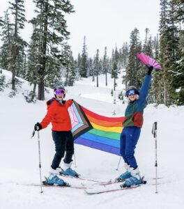 two alpine skiiers dressed in bright colors holding a Progress Pride Flag in the snow