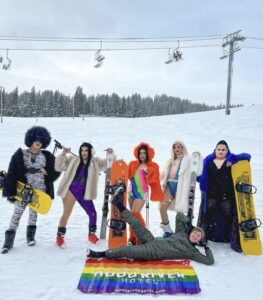 A group of drag queen skiiers with one man laying on the ground in front of them posing with a Pride Flag in the snowy mountians
